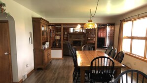 A dining area with a wooden table and chairs, a chandelier, and a kitchen in the background.