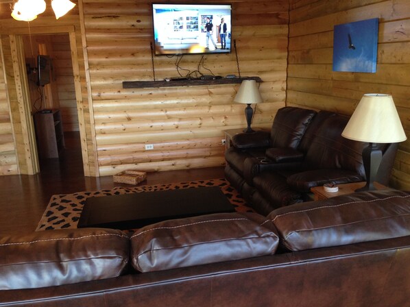 A log cabin interior with a leather sofa, a television mounted on the wall, and a lamp on a side table.