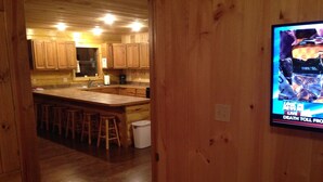Un interior de madera con zona de cocina que incluye armarios de madera, una isla central con taburetes y un televisor que muestra un noticiero.
