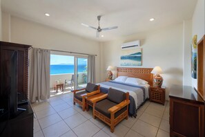 A hotel room with a bed, a ceiling fan, a TV, a balcony with a view of the ocean, and a wooden side table with a lamp.