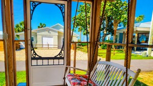 A wooden porch with a swing chair and a view of a house and palm trees. -  Coastal Cottage across from the beach, pet friendly, fenced yard (Port Saint Joe)
