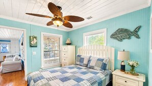 A bedroom with a wooden ceiling fan, a bed with a blue and white checkered bedspread, a white nightstand with a lamp, and a fish-shaped wall decoration. -  Coastal Cottage across from the beach, pet friendly, fenced yard (Port Saint Joe)