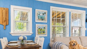 A dining area with a wooden table set for four, a beige sofa with a dog, and two windows with shutters. -  Coastal Cottage across from the beach, pet friendly, fenced yard (Port Saint Joe)