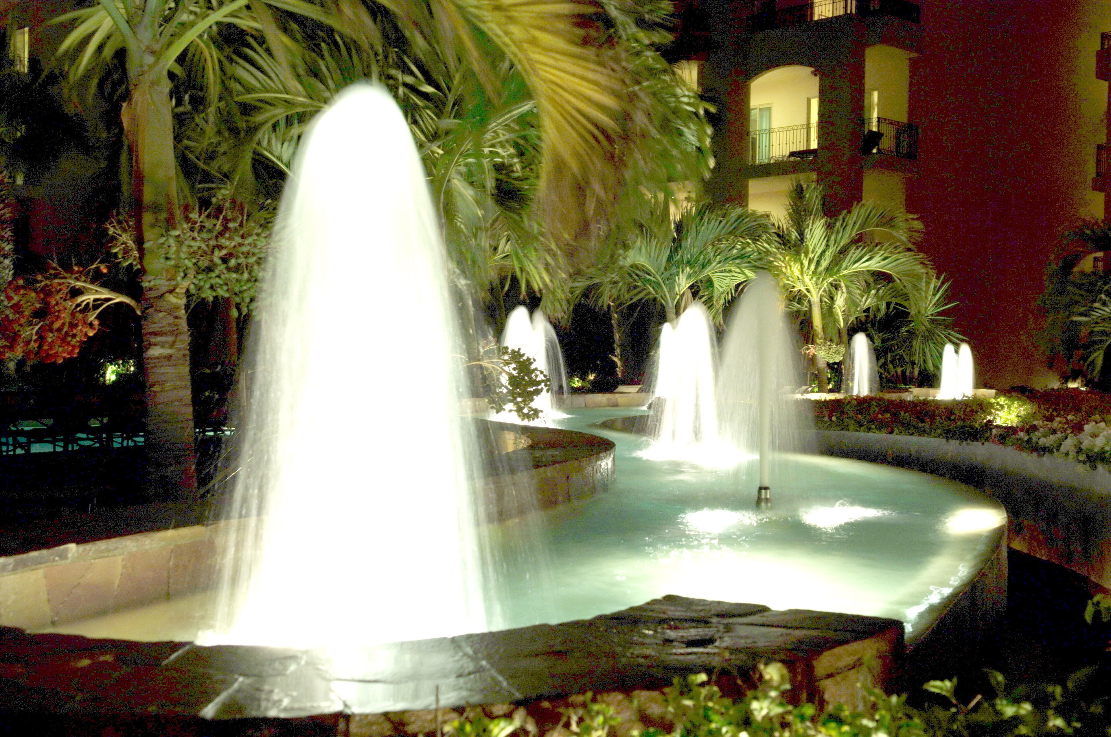 A nighttime scene of a fountain with multiple water jets, surrounded by palm trees and a building with balconies in the background.