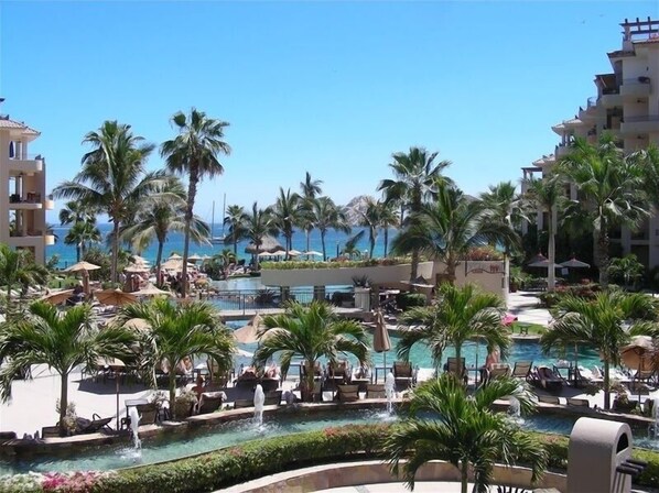 Relax by the central pool surrounded by palm trees and lounge areas, with a scenic ocean view.