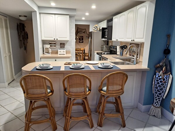 A modern kitchen with white cabinets, a central island with bar stools, and a dining area with a table and chairs. - Gorgeous Sunsets from Your Balcony in Moon Bay/Walk to nearby restaurants (Key Largo)