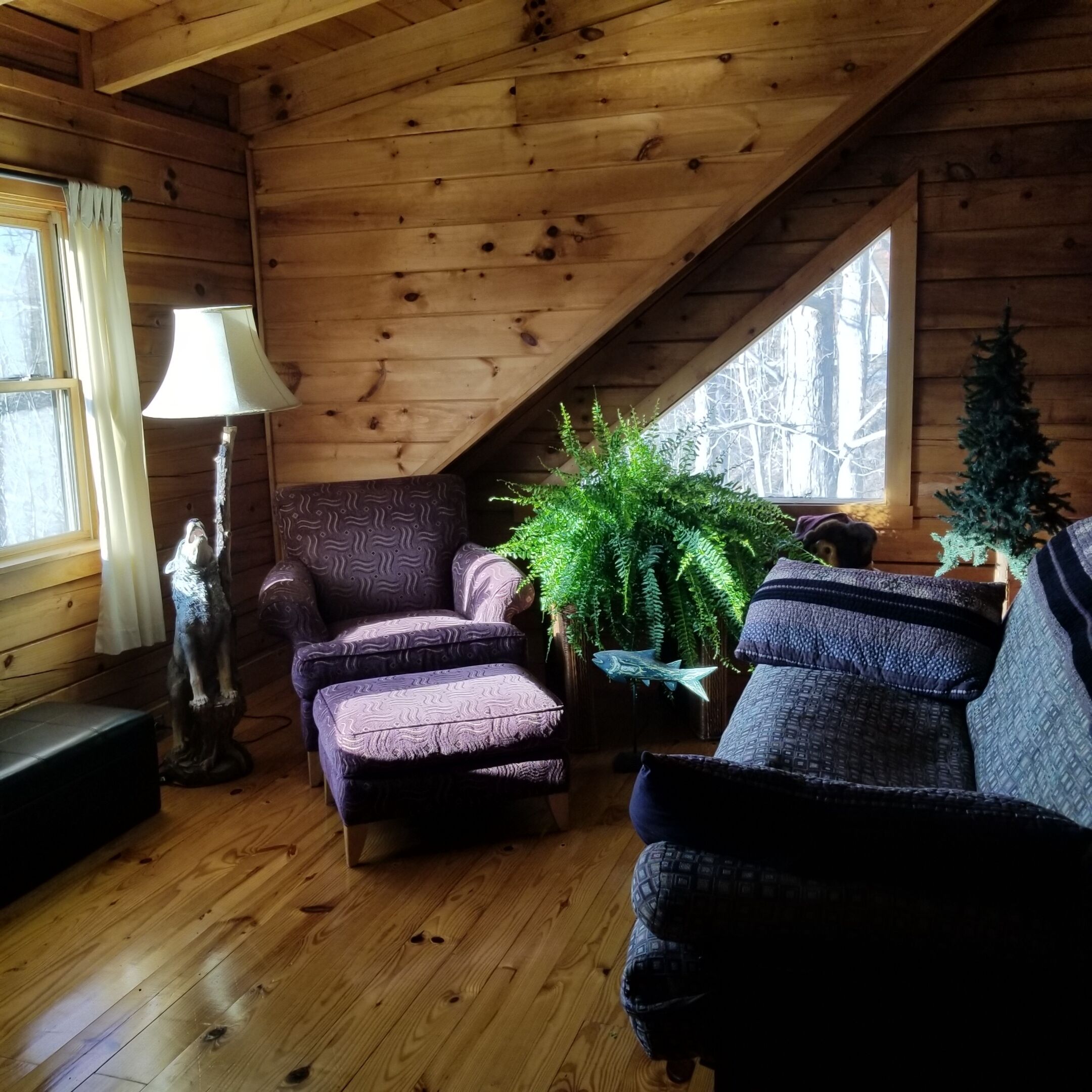 A cozy cabin interior with wooden walls, a sloped ceiling, a small table, two patterned armchairs, a floor lamp, and a Christmas tree.