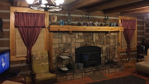 A cozy log cabin interior with a stone fireplace, wooden beams, a chandelier, and a television.