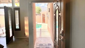 Un couloir mène à un espace piscine agrémenté d’un paillasson « Bienvenue », de portes en bois et offrant une vue sur la piscine et les montagnes.