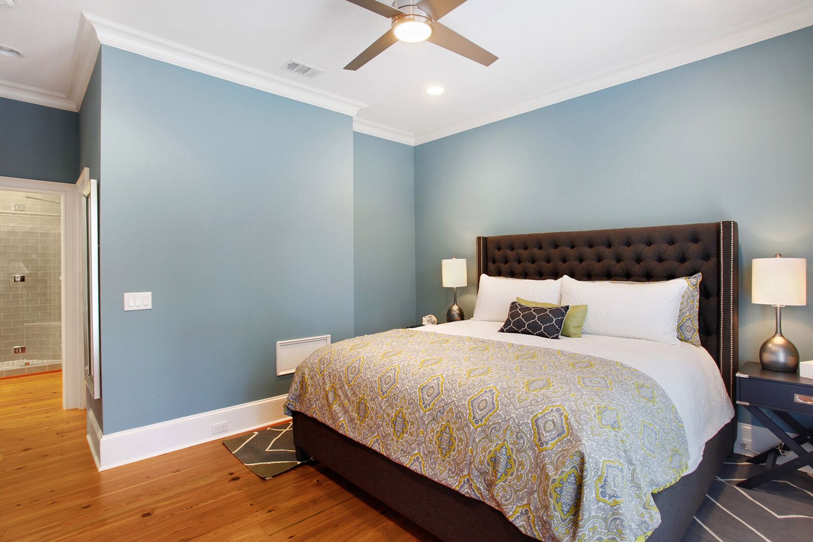 Quarto com cabeceira estofada, colcha estampada, duas luminárias de mesa e piso de madeira.