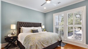 A bedroom with a tufted headboard, a patterned bedspread, two bedside lamps, and a window with blinds. - Wright Square Loft: Amazing Unit A in Downtown Savannah With Great Views! (Savannah)