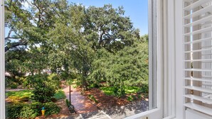 A view through a window with white blinds, featuring a well-maintained garden with various trees and shrubs, a paved path, and a yellow fire hydrant. - Wright Square Loft: Amazing Unit A in Downtown Savannah With Great Views! (Savannah)