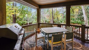 A covered outdoor area with a grill, a table, and chairs, surrounded by a wooden railing and a view of a forest. - Perfect Family Home w/ Pool & Lagoon Views (Hilton Head Island)
