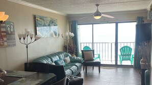 A living room with a green sofa, a dining table with chairs, a ceiling fan, and a view of the ocean through a sliding door. - Beautiful  2 Bedroom Vacation Condo in Galveston - Great Gulf Views   (Galveston)