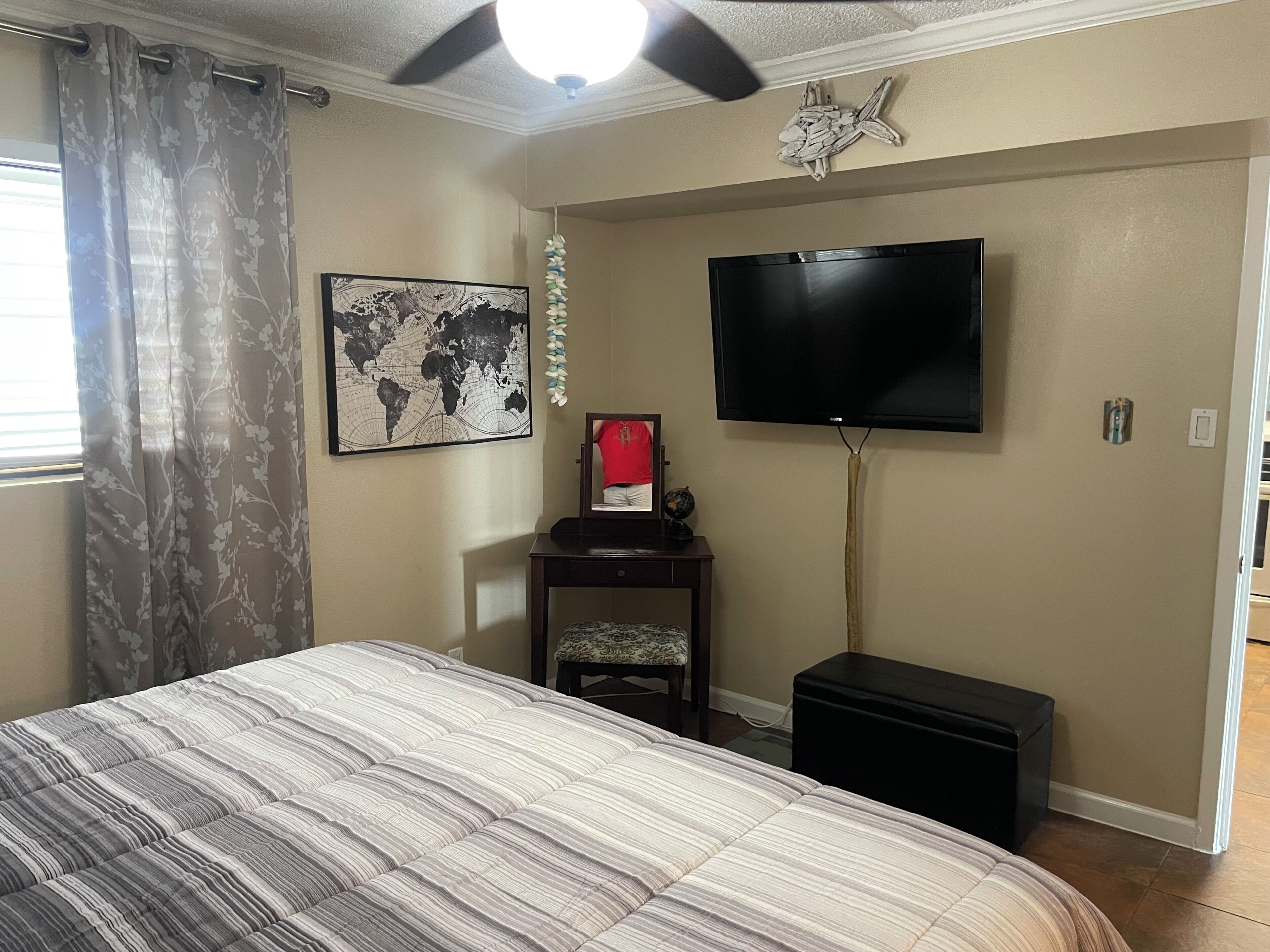A bedroom with a bed, a flat-screen TV mounted on the wall, a world map artwork, a globe, and a ceiling fan.