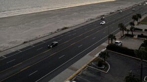 A coastal road with a beach on one side and a parking lot with palm trees on the other. - Beautiful  2 Bedroom Vacation Condo in Galveston - Great Gulf Views   (Galveston)