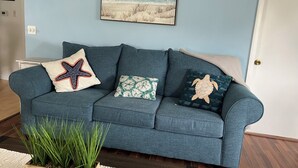 Ein blaues Sofa mit drei Zierkissen, ein Holzpflanzentisch und ein gerahmtes Strandbild an der Wand.