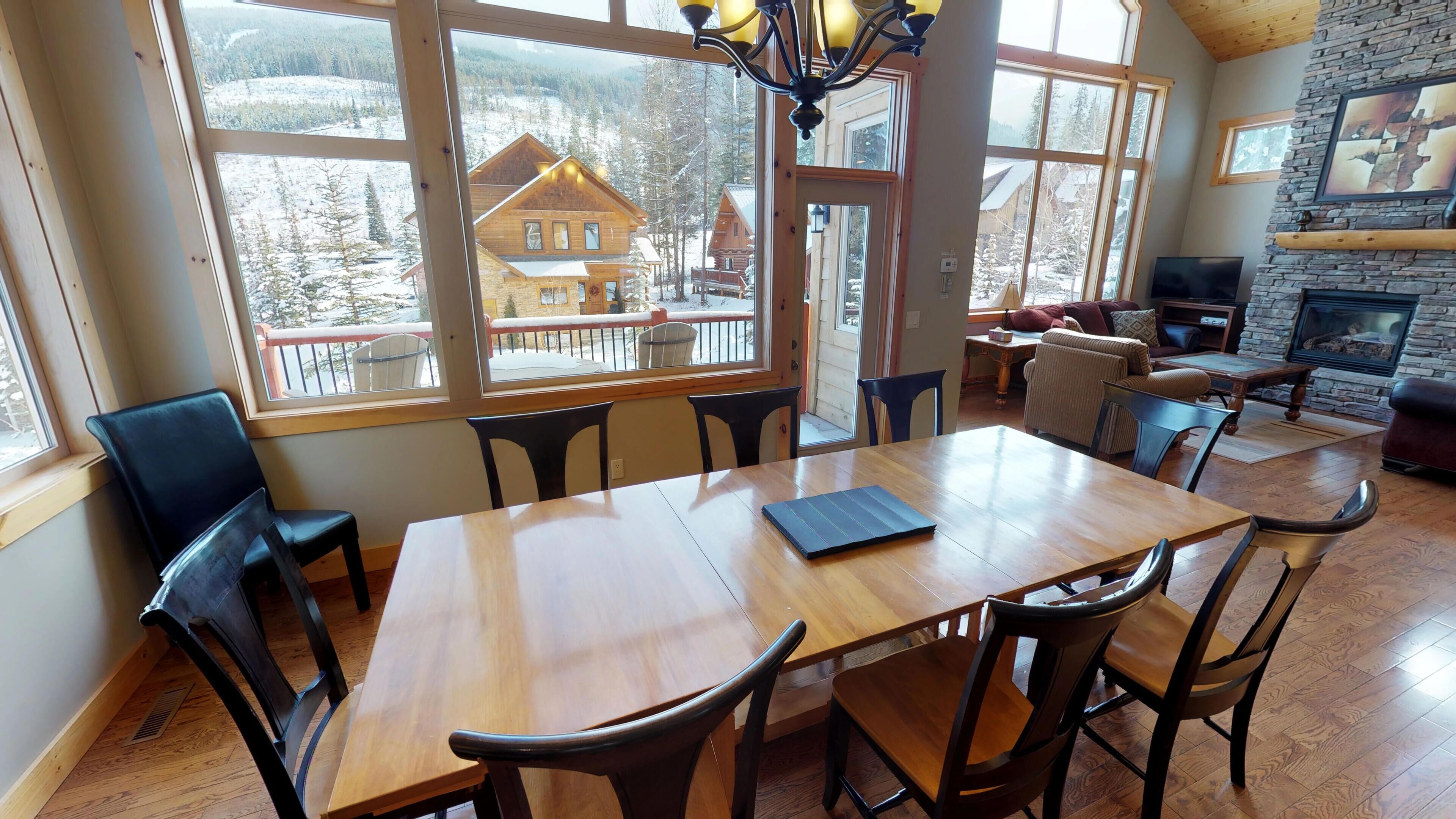 Een eetruimte met een houten tafel en stoelen, grote ramen met uitzicht op een besneeuwde berg en houten chalets, een stenen open haard en een knus zithoekje met een bank en fauteuils.