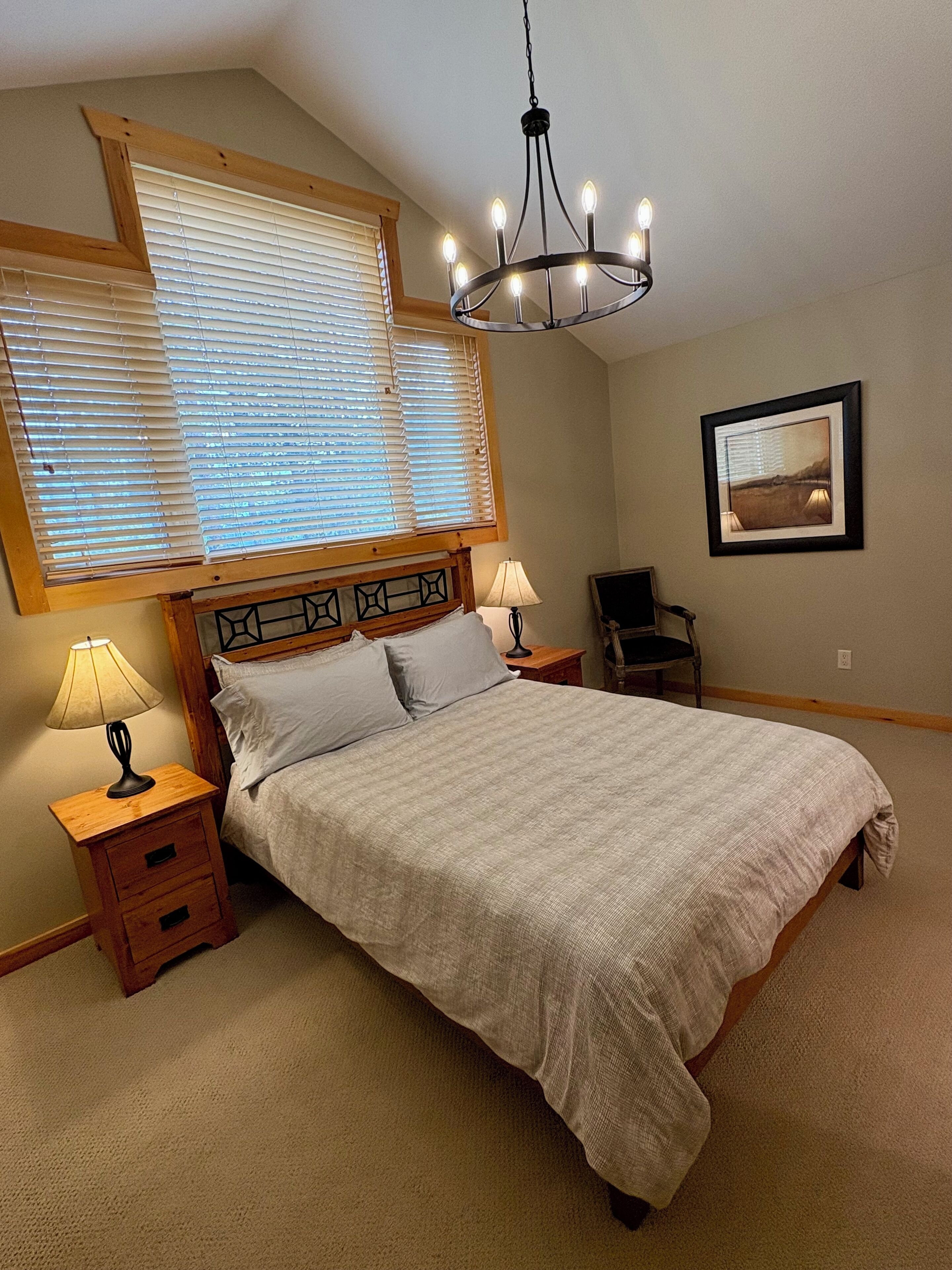A bedroom with a large bed, two bedside tables with lamps, a wooden headboard, a chandelier, a window with blinds, a framed picture on the wall, and a chair.