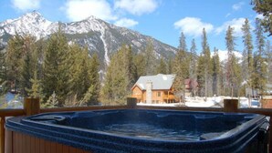 A hot tub with a wooden deck, a mountain backdrop, and a lodge with a red roof. - Spectacular Mountain Views | Private Hot Tub | Fire Pit | Central A/C (Panorama)