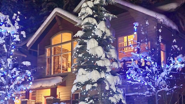 A snow-covered landscape with a wooden house featuring large windows and a visible fireplace. - Spectacular Mountain Views | Private Hot Tub | Fire Pit | Central A/C (Panorama)