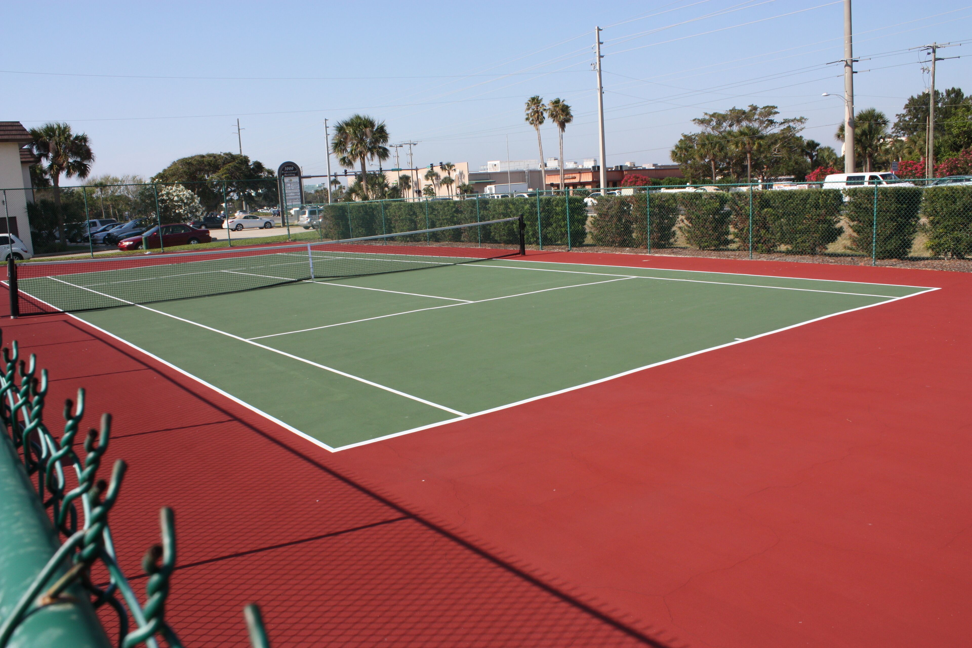A tennis court with a net, surrounded by a green fence and palm trees.