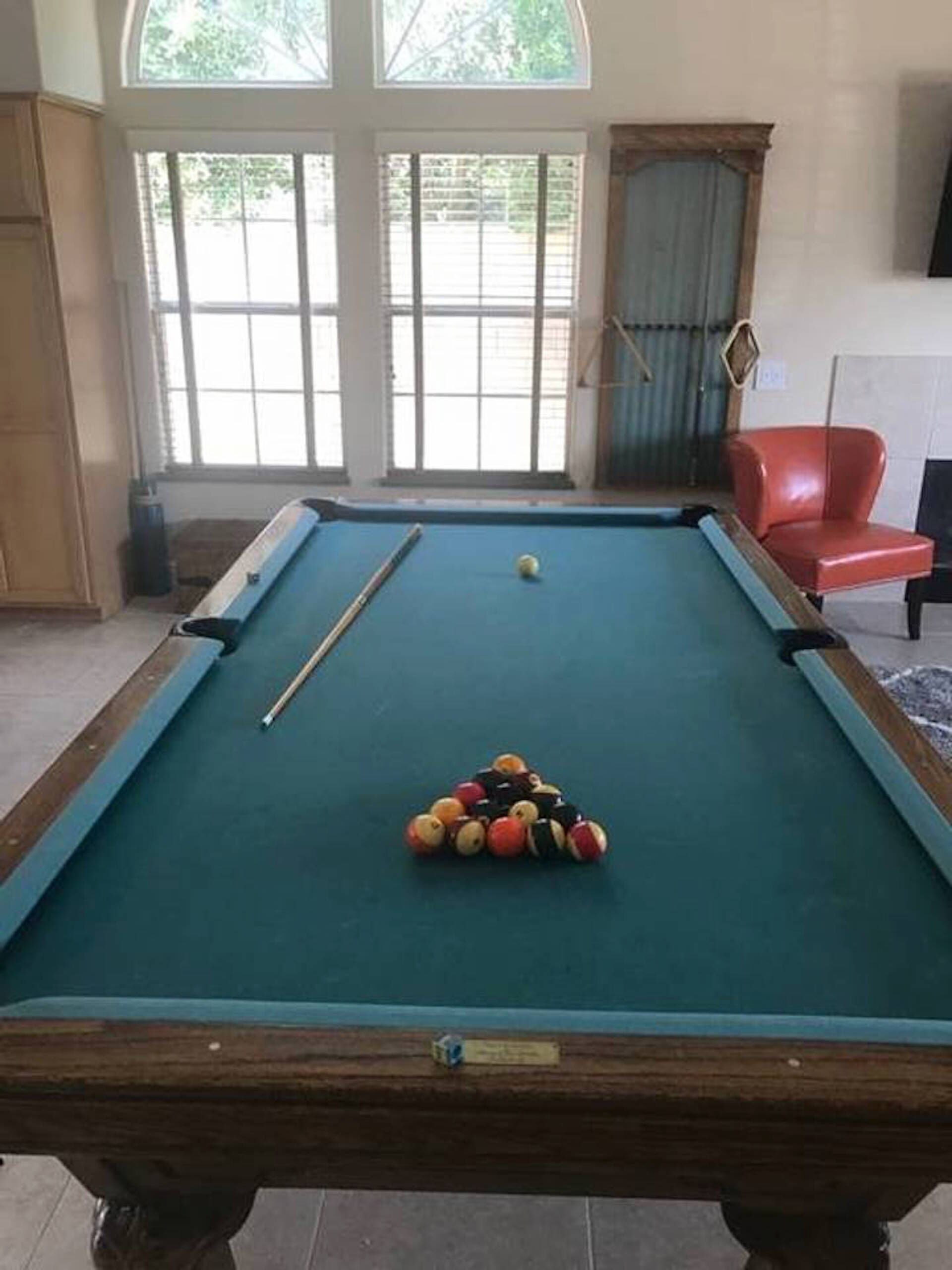 A pool table with balls, a pool cue, and a red chair in a room with large windows and a view of greenery outside.