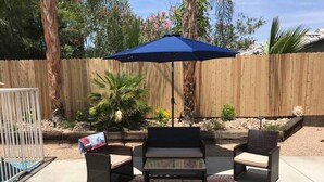 A patio with two black wicker chairs, a dark brown rug, and a blue umbrella. - BRAND NEW, PRISTINE HAVEN WITH SALT WATER  POOLS (Palm Springs)