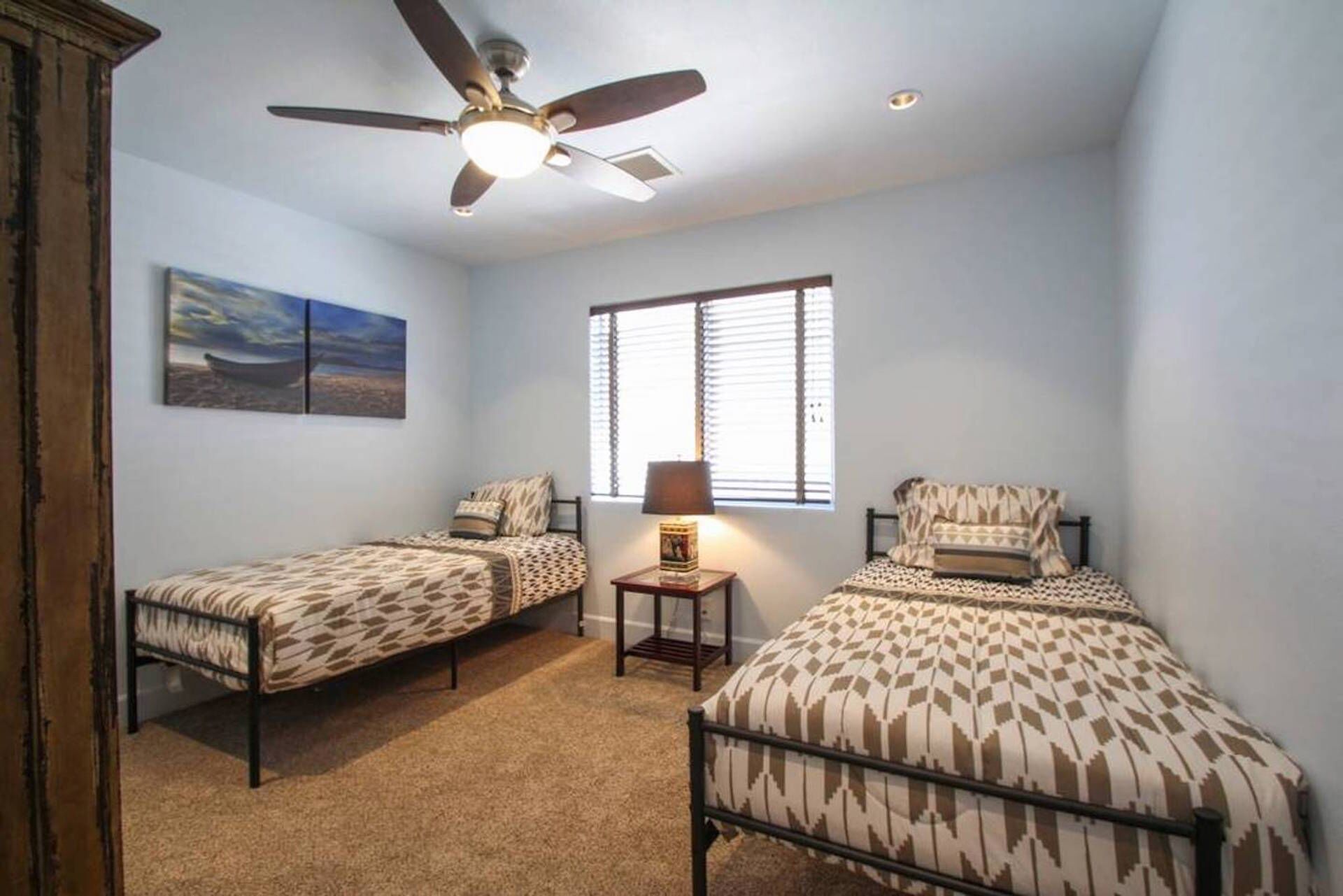 A bedroom with two single beds, a ceiling fan, a window with blinds, and a painting on the wall.