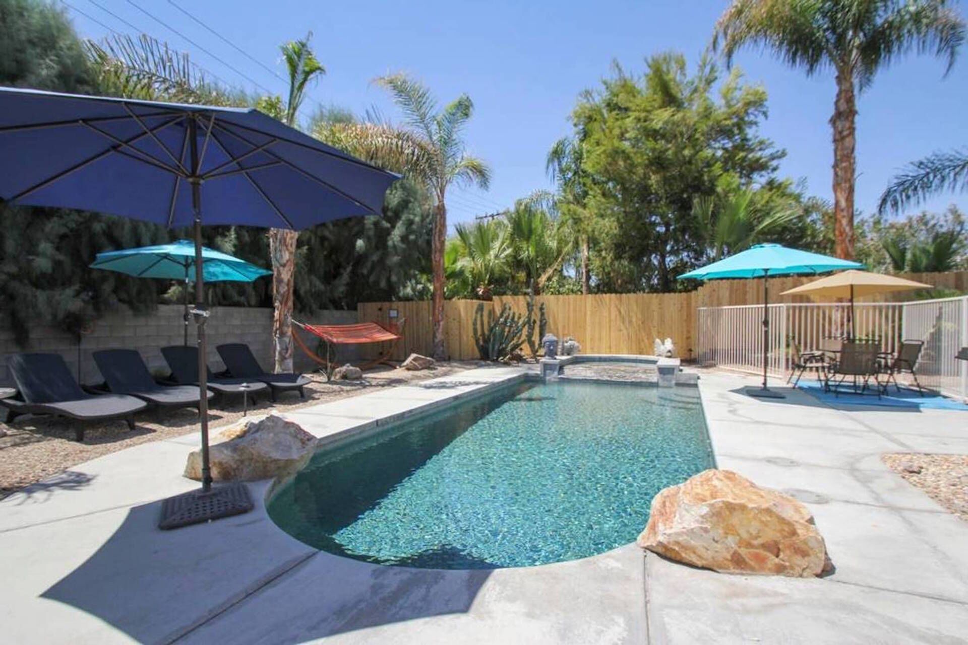 A rectangular swimming pool with clear water, surrounded by lounge chairs and umbrellas, and a wooden fence in the background.