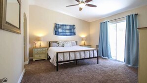 A bedroom with a bed, two bedside tables, a ceiling fan, a wall tapestry, and a large window with curtains. - BRAND NEW, PRISTINE HAVEN WITH SALT WATER  POOLS (Palm Springs)