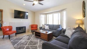 A living room with a fireplace, a ceiling fan, a flat-screen TV, and a large window with blinds. - BRAND NEW, PRISTINE HAVEN WITH SALT WATER  POOLS (Palm Springs)