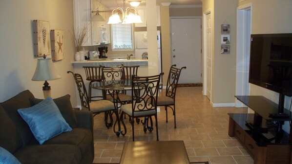 A living room with a brown sofa, a coffee table, and a TV stand. There is a dining area with a table and chairs, and a kitchen in the background. - Newly Renovated Condo in Beautiful Beach Front Resort (Hilton Head Island)