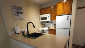 Cozinha compacta com armários de madeira, bancada branca, pia, fogão, micro-ondas e geladeira.