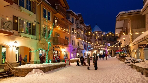 Rua coberta de neve com edifícios de ambos os lados, iluminada por luzes de Natal.