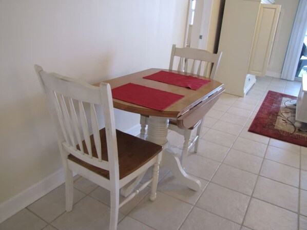 A dining area with a wooden table and white chairs, a red placemat, and a tiled floor.