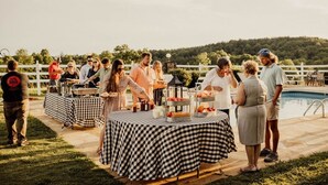 Un grupo de personas en un evento al aire libre, con mesas a cuadros, una piscina y una cerca blanca al fondo.