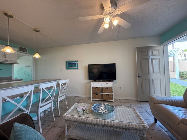 A living room with a TV, a ceiling fan, a dining area with chairs, and a kitchen in the background. - First Floor/Directly on the Beach/End Unit (Fort Walton Beach)