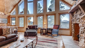 A spacious living room with large windows, a stone fireplace, and a cozy seating area with a rocking chair and a sofa. - Log Home, Hot Tub & Pool-Table Game Room - 25 mi to Breckenridge (Fairplay)