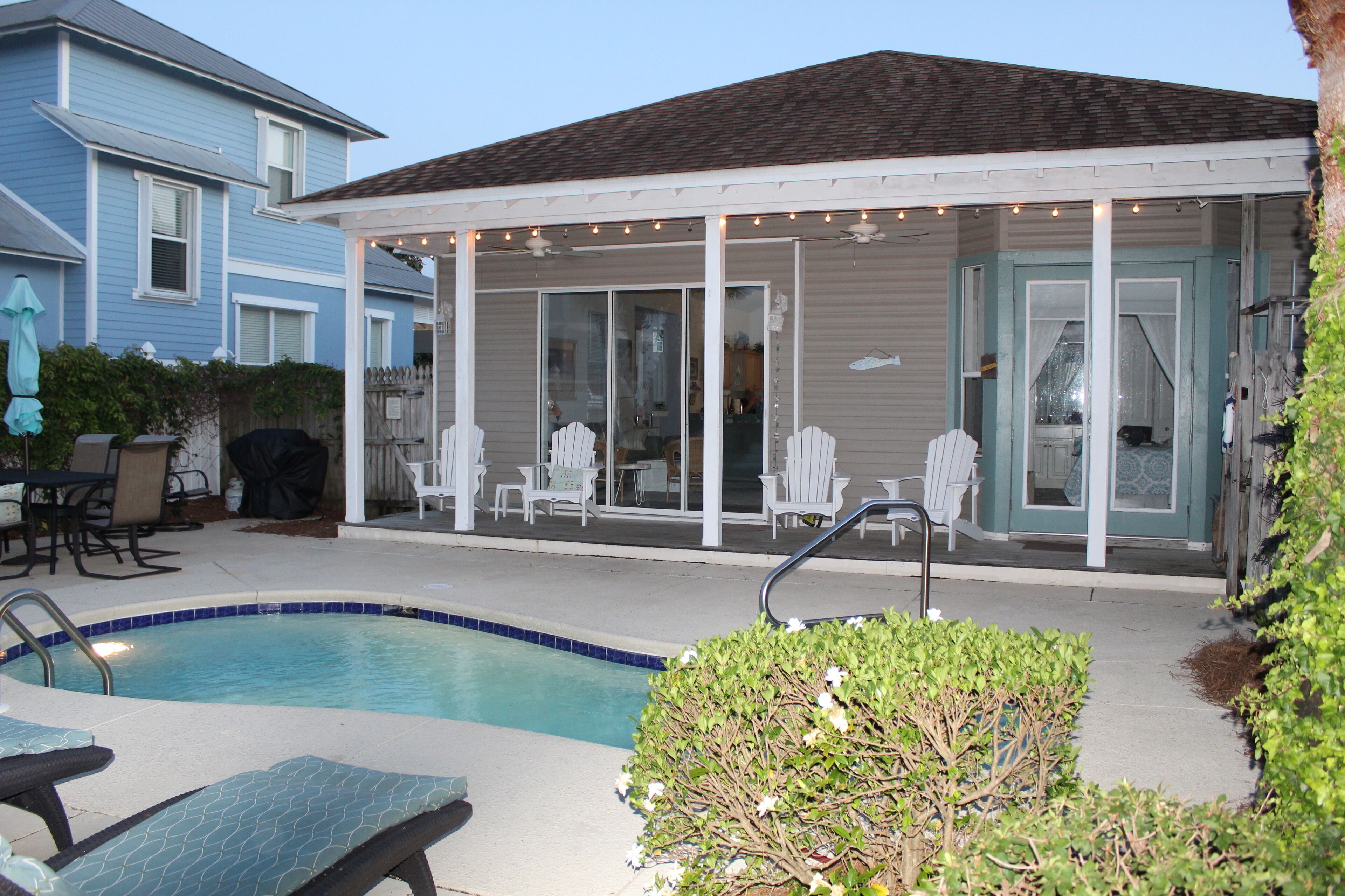 A covered patio with a pool, white chairs, and a small table.