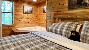 Een slaapkamer in een houten blokhut met een bed, hoofdbord, raam en een schilderij van een beer.