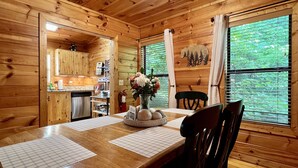 Interieur van een houten chalet met een eettafel, stoelen, een vaas met bloemen, een keukenhoek en grote ramen met gordijnen.