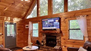Een gezellig chalet interieur met een stenen openhaard, leren banken, een televisie boven de openhaard en grote ramen met gordijnen.