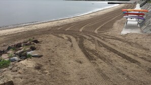 Una playa con kayaks alineados sobre una estructura de concreto, un sendero arenoso que lleva al agua, y una vista distante de casas y una bandera.