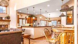 A spacious living area with a fireplace, a dining table with chairs, and a kitchen area with wooden cabinets and hanging pendant lights.