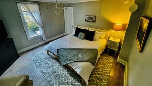 A bedroom with a bed, a chandelier, a nightstand, a lamp, a dresser, and a window with curtains. - Peaceful Cottage Retreat Near Nashville! (Ashland City)
