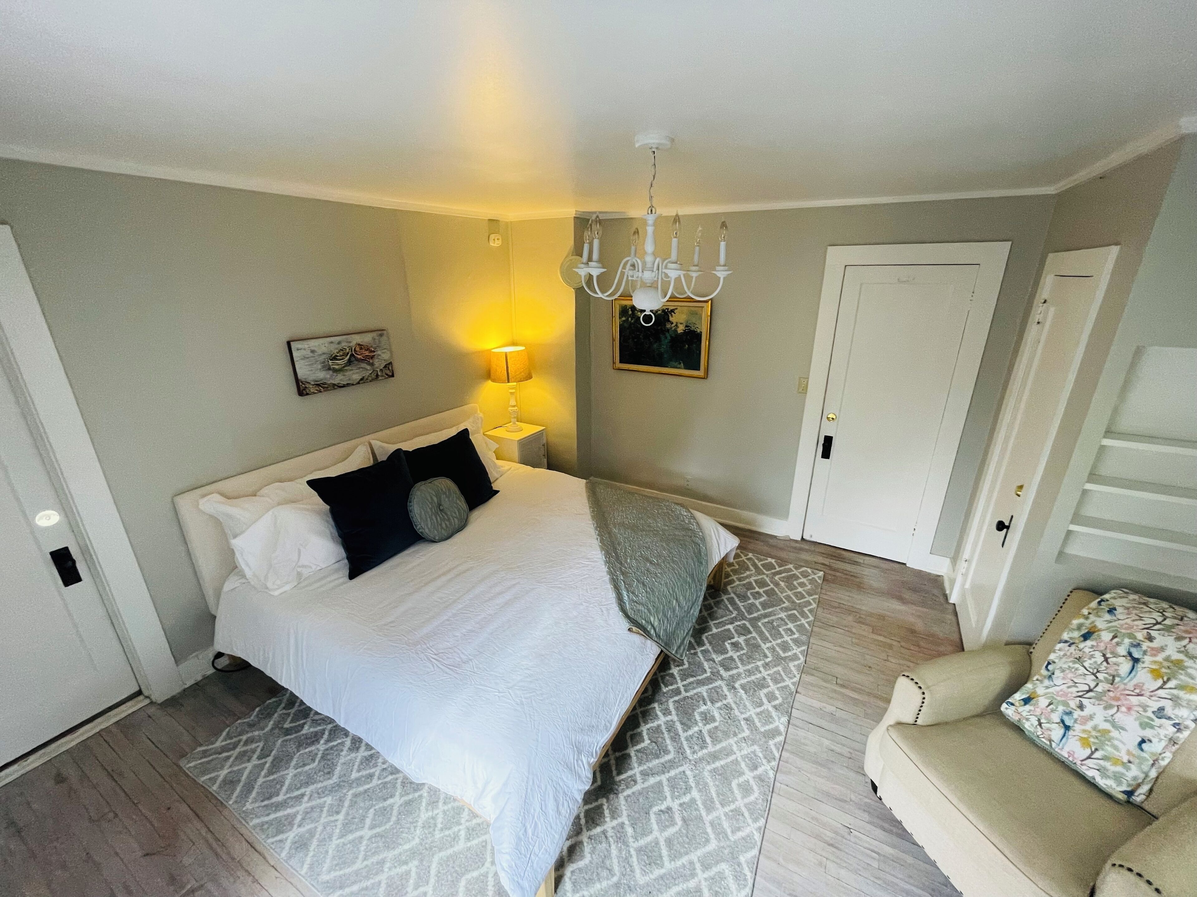 Ein Schlafzimmer mit einem Bett, einem Nachttisch mit Lampe, einer Chandelierleuchtre, einer Tür und einem gemusterten Teppich.
