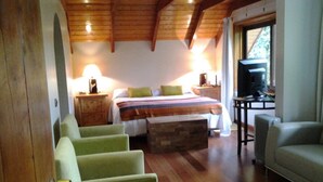 A cozy room with a wooden ceiling, a bed with a striped blanket, a green armchair, a wooden chest, and a television on a table.