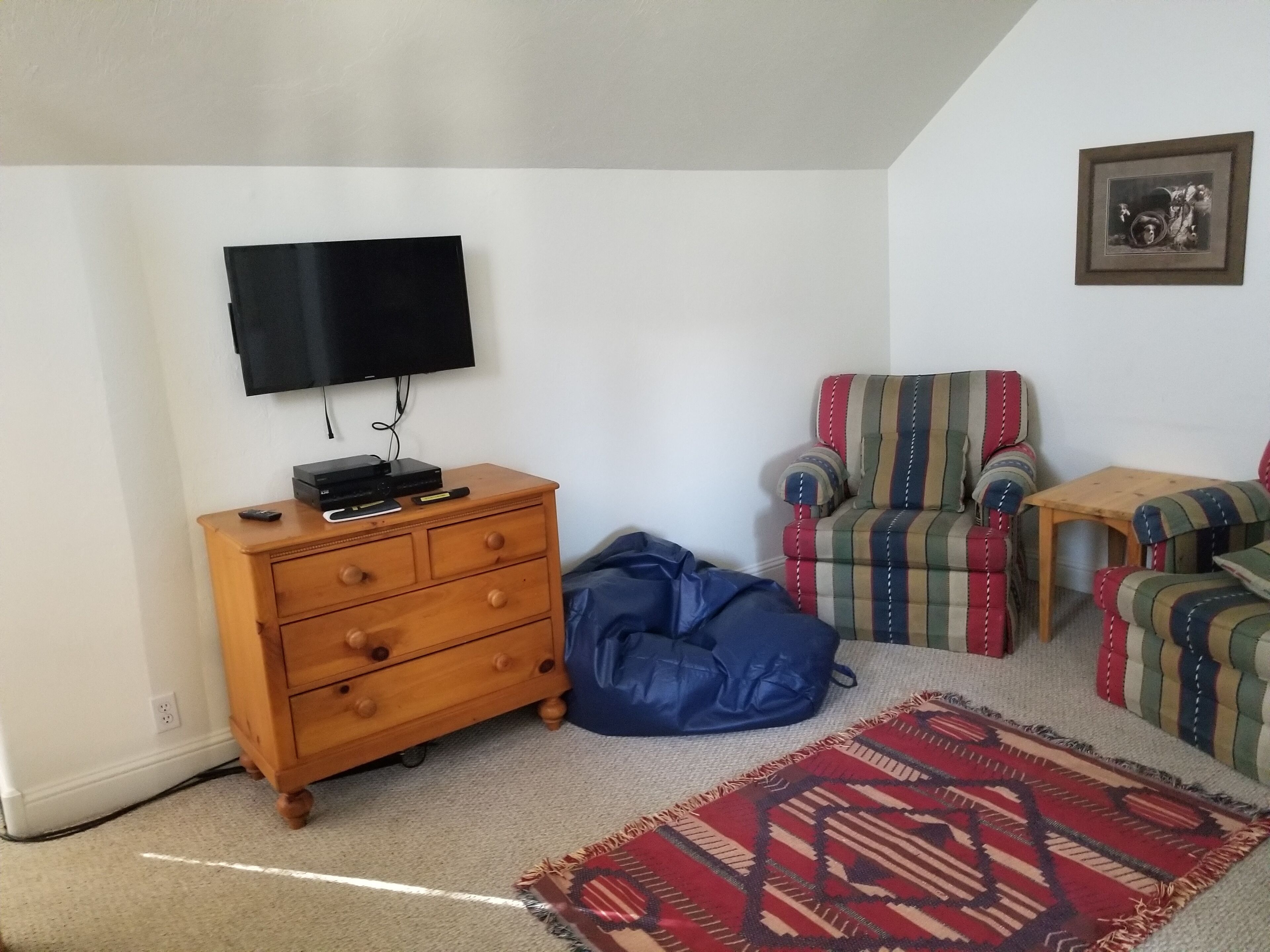 A room with a wooden dresser, a television mounted on the wall, a striped armchair, a small wooden table, and a patterned rug.