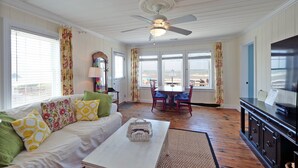 En stue med ventilator i loftet, hvid sofa med mønstrede puder, sofabord i træ, samt spiseområde med bord og stole.